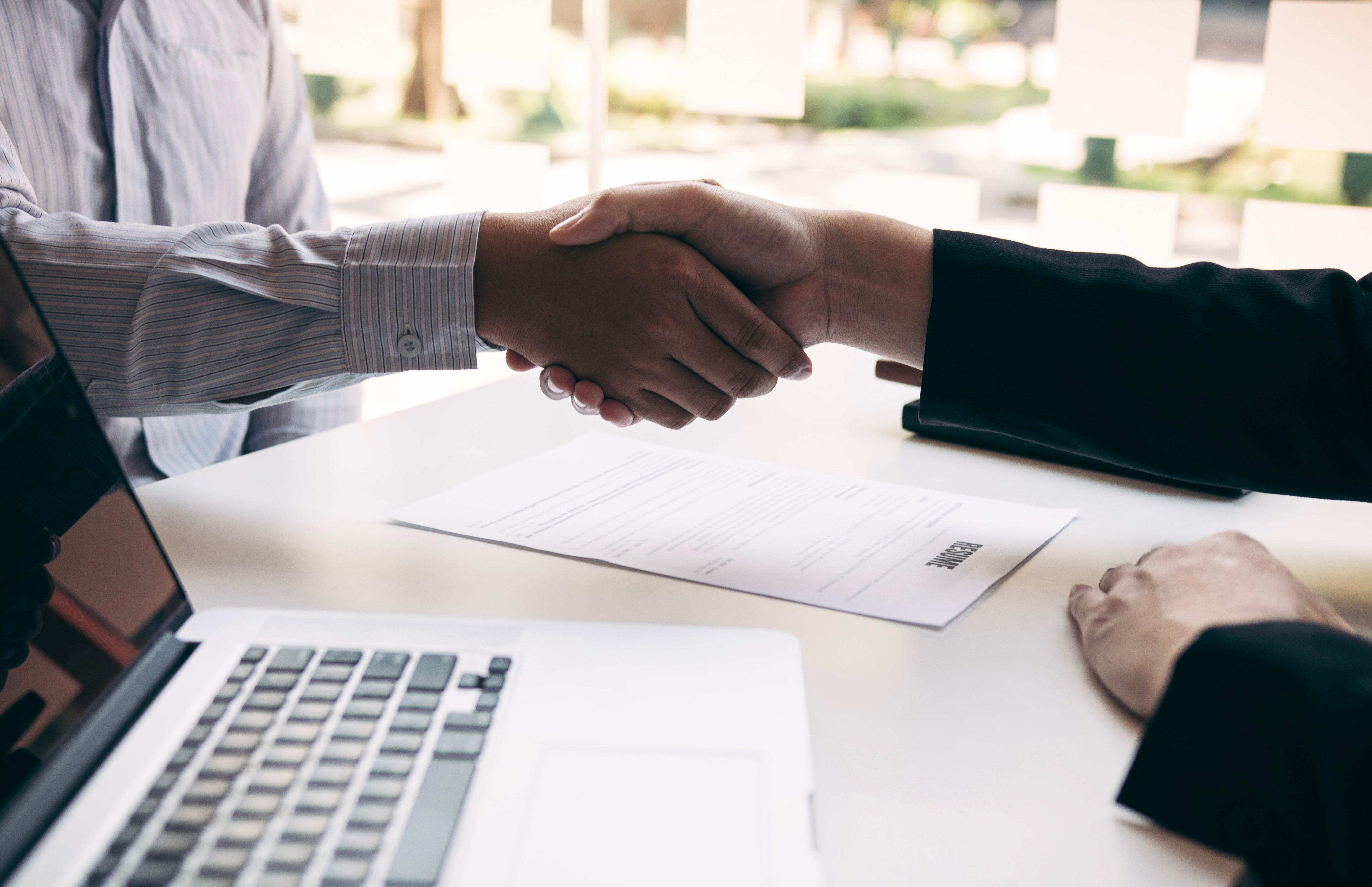 Image of a handshake over a desk with a piece of paper. and laptop